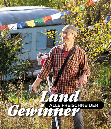Mann schneidet  mit einem Freischneider Gestrüpp auf einem verwilderten Grundstück mit einem Bauwagen im Hintergrund